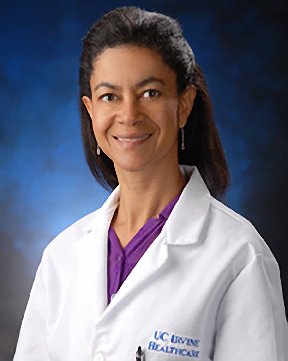 Headshot of Joan Campbell wearing a white doctors coat. She is an African American woman with shoulder length black hair, She is wearing a purple button down shirt under her white coat and she is posing in front of a blue background. 