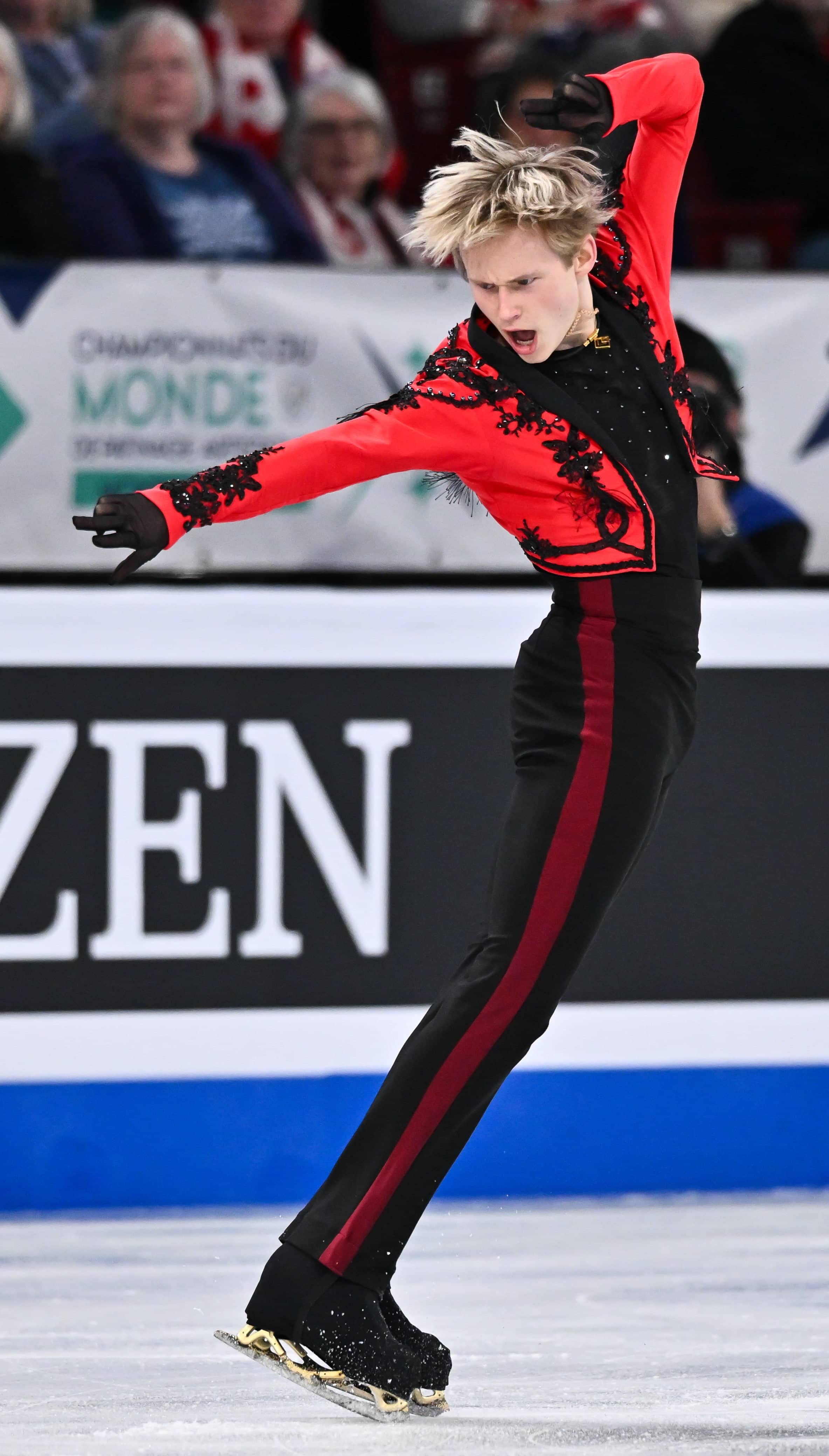 Ilia Malinin strikes a pose with one arm above his head and the other pointing forward. He is wearing a red and black long sleeve shirt and black pants with a red stripe down the side