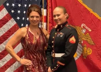 Dressed in her formal U.S. Marine uniform, Stephanie Kazior stands next to wife, Jill Ahlbrecht, who is wearing a sleeveless red dress. They are standing in front of an American flag and a U.S. Marine Corps flag.