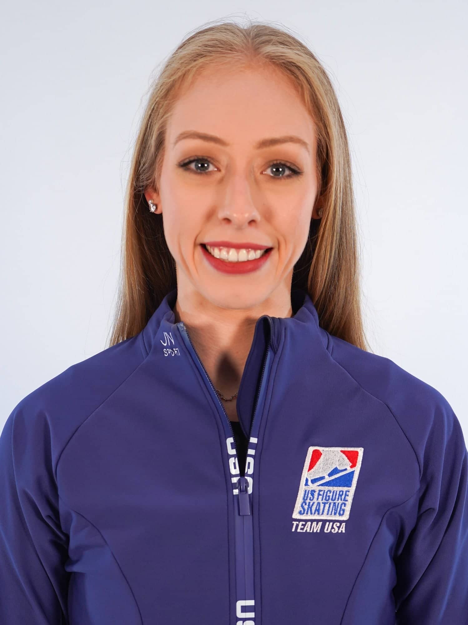  Bradie Tennell headshot. She stands in front of a white backdrop, facing the camera straight on. She has long blonde hair and is wearing a a blue Team USA jacket and is smiling.