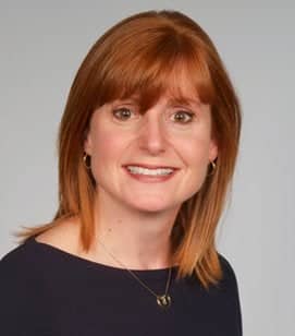 Headshot of a Caucasian woman smiling. She has shoulder length red hair with bangs. She is wearing a black shirt and a dainty necklace. 